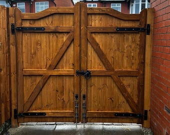 May include: A pair of wooden double gates with black metal hinges and latches. The gates are closed and are in front of a brick wall.