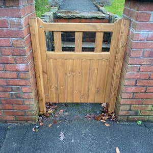 May include: A wooden gate with a slatted design, painted in a light brown colour, is set between two brick pillars. The gate is closed and there are some fallen leaves on the ground in front of it.