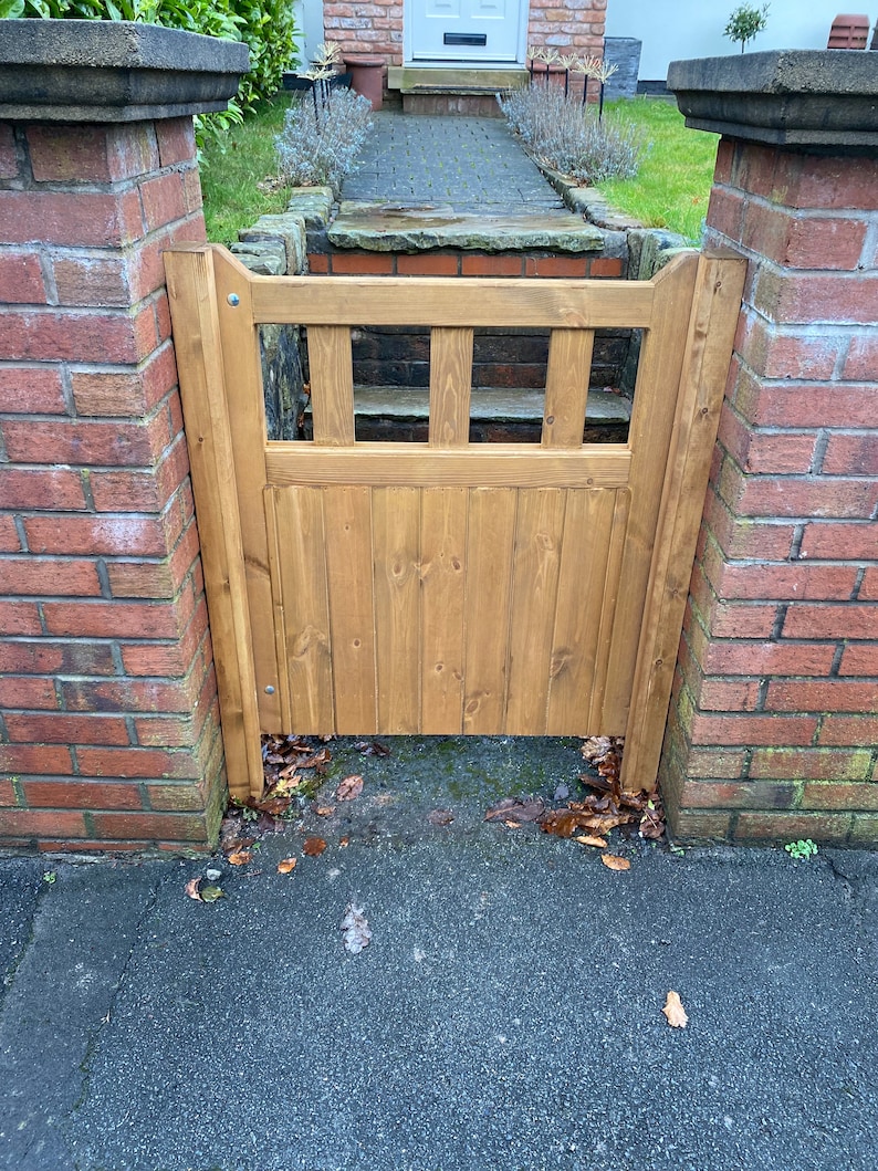 May include: A wooden gate with a slatted design, painted in a light brown colour, is set between two brick pillars. The gate is closed and there are some fallen leaves on the ground in front of it.