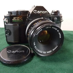 Könnte beinhalten: Eine schwarze Canon AE-1 Program Kamera mit einem 50mm Objektiv und Objektivdeckel. Das Objektiv ist mit "LENS MADE IN JAPAN" gekennzeichnet. Der Kamerabody hat den Text "AE-1 PROGRAM" in Grün. Der Objektivdeckel hat das Canon Logo.
