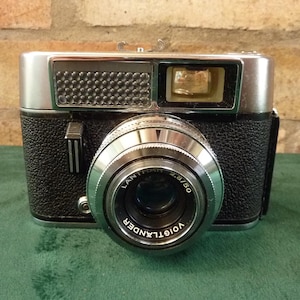 May include: A vintage Voigtlander camera with a silver lens and black textured body. The lens is marked 'LANTHAR 2.8/50'. The camera has a silver top with a viewfinder and flash. The camera is on a green surface.