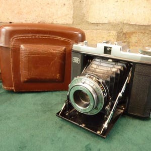 May include: A vintage black and silver camera with a brown leather case. The camera has a bellows system and a lens with a silver ring. The case has a strap.