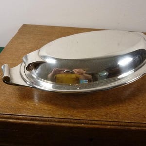 May include: A polished, oval silver serving dish with a lid. The dish features two handles and a reflective surface. It is placed on a wooden surface.