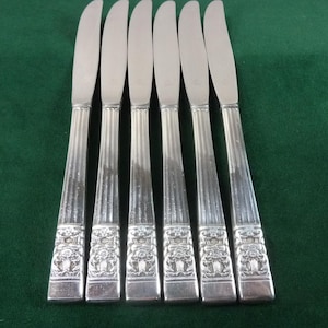 Puede incluir: Seis cuchillos de mantequilla plateados con un diseño floral decorativo en los mangos. Los cuchillos están dispuestos en fila sobre una superficie verde oscuro. Las hojas son lisas y curvas.
