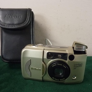 May include: A gold Nikon Lite-Touch Zoom 70W AF camera with a black lens and a black leather case. The camera has a pop-up flash. The Nikon logo is visible on the camera and case.