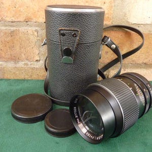 Puede incluir: Un objetivo de cámara negro con un anillo plateado y una funda de transporte de cuero negro. El objetivo tiene un anillo negro y plateado con el texto "T2.8 1:135 No. 377328" impreso en él.