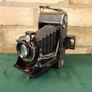 May include: A vintage black folding camera with a bellows system and a lens with a silver ring. The camera has a silver lever on the side and a small viewfinder on the top.