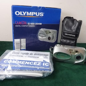 Könnte beinhalten: Eine silber-grüne Olympus Camedia C-450 Zoom Digitalkamera mit schwarzer Ledertasche und Bedienungsanleitung. Der Markenname Olympus ist auf der Box und der Tasche zu sehen.