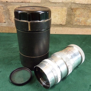 Può includere: Un obiettivo per fotocamera argentato con una custodia in pelle nera. L'obiettivo ha un corpo argentato con segni neri e un anello di gomma nero attorno alla parte anteriore. La custodia è in pelle nera con un bordo dorato.