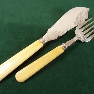 Puede incluir: Un juego de cuchillo de mantequilla y tenedor con mangos de color crema y hojas de metal plateado. El cuchillo tiene una hoja decorativa y curvada con patrones florales grabados. El tenedor tiene cuatro púas. El juego se muestra sobre una superficie verde.