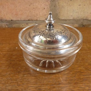 May include: A clear glass jar with a silver lid. The lid features a decorative floral pattern and a small, pointed finial. The jar is on a wooden surface.