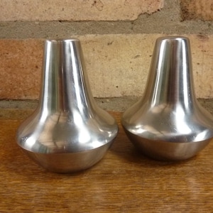 Puede incluir: Dos saleros y pimenteros plateados y brillantes a juego. Cada uno tiene una base bulbosa y una parte superior cónica. Se encuentran sobre una superficie de madera contra un fondo de pared de ladrillo. Los saleros son de metal.