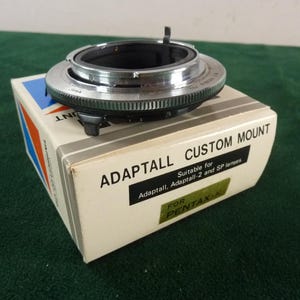 Op de afbeelding: Een zilver- en zwartkleurige metalen cameralensvatting-adapter op een witte kartonnen doos. De doos is bedrukt met de tekst ADAPTALL CUSTOM MOUNT, Suitable for Adaptall, Adaptall-2 and SP lenses en FOR PENTAX-K op de voorzijde.