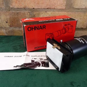 Può includere: Un duplicatore di diapositive Ohnar Zoom nero con uno schermo bianco e un obiettivo nero. Il duplicatore è su una superficie verde. La scatola del duplicatore è rossa e dice "Ohnar Zoom Slide Duplicator".