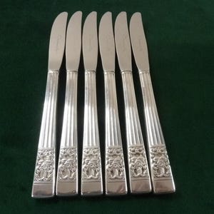 Peut inclure: Six couteaux à beurre en argent. Les manches présentent un design côtelé et un motif floral détaillé à la base. Les lames sont incurvées et pointues. Les couteaux sont disposés sur une surface vert foncé.