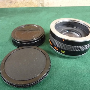 May include: A camera lens adapter with two black lens caps. The adapter is black and silver with the text "MC4 OM". The caps are round and black. The items are on a green surface.