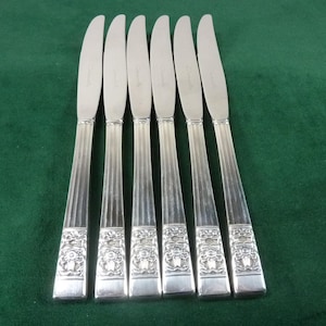 Puede incluir: Juego de seis cuchillos de mantequilla plateados. Cada cuchillo presenta una hoja curva y un mango con estrías verticales y un diseño ornamental decorativo en la base. Los cuchillos están sobre una superficie verde.