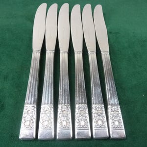 Puede incluir: Seis cuchillos de mantequilla plateados sobre una superficie verde. Los cuchillos tienen una hoja curva y un mango decorativo con líneas verticales y un diseño floral. Las hojas tienen una ligera curvatura.