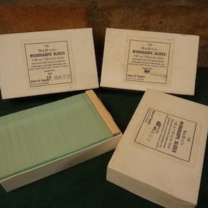 Könnte beinhalten: Vier Kartons mit Vintage-Mikroskopobjektträgern. Jeder Karton ist mit Text beschriftet, darunter "100 Mikroskop-Objektträger 76 x 25 mm" und "1:00 bis 1:20 mm dick". Ein Karton ist geöffnet und zeigt einen Stapel grüner Glasobjektträger.