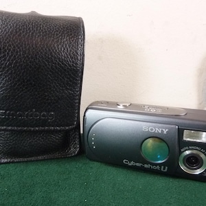 May include: A black leather camera case with the word "smartbag" embossed on the front. Next to it is a gray Sony Cyber-shot U digital camera with a lens, flash, and the words "Cyber-shot U" printed on the front.