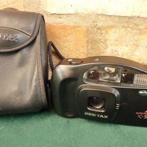 Puede incluir: Una cámara Pentax K-50 autofocus negra con una funda de cuero negra. La cámara tiene un flash incorporado y una tapa de lente.