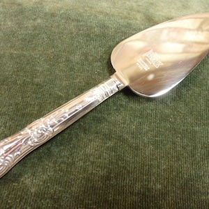 May include: Silver-colored pie server with a decorative handle. The server has a wide, triangular blade and a handle with an ornate, floral design. The server is made of a shiny, reflective metal.