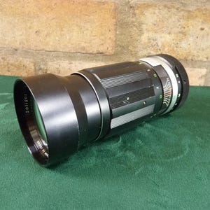 Puede incluir: Un objetivo de cámara Soligor negro con parasol, sobre una superficie verde. El objetivo tiene un anillo plateado con marcas de medición y un cuerpo negro con detalles acanalados. El parasol lleva el nombre de la marca Soligor.