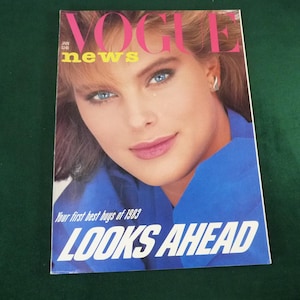 May include: A vintage January 1983 Vogue News magazine cover featuring a close-up portrait of a person with blue eyes and brown hair wearing a bright blue garment. The text reads VOGUE, news, Your first best buys of 1983, and LOOKS AHEAD.
