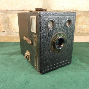 Puede incluir: Una cámara de caja vintage negra con exterior de cuero y una lente marcada con "Popular Brownie Takes No. 120 Kodak Film".