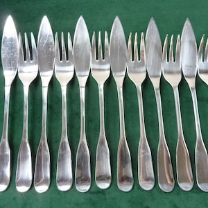 May include: A set of silver-colored flatware, including knives and forks, arranged on a green surface. The knives have a pointed blade, and the forks have three tines. The flatware is likely for a formal dining setting.