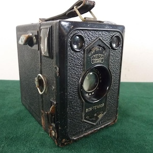 May include: A black Zeiss Ikon Box-Tengor camera with a textured finish, a leather carrying strap on top, and a central lens. The front plate features the brand name, the model name, and a small metal lever for aperture adjustments.