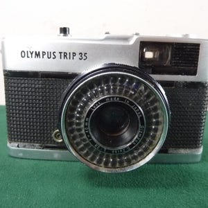 Op de afbeelding: Olympus Trip 35 camera met een zilveren bovenkant en zwarte getextureerde behuizing. De lens is zwart met zilveren accenten en de tekst "Lens made in Japan" is zichtbaar. De camera staat op een groene ondergrond.
