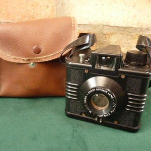 Puede incluir: Una cámara vintage negra Envoy Optimax con una funda de cuero marrón. La cámara tiene un objetivo con el texto "Envoy Optimax Lens" y "Camera - 127 - Portraits - Pull Out - 127 -  " impreso en ella.
