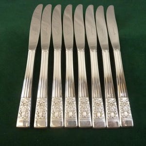 Puede incluir: Un juego de ocho cuchillos plateados con mangos ornamentados y hojas estriadas. Los mangos presentan un diseño floral. Los cuchillos están dispuestos sobre una superficie verde oscuro, mostrando su elegante diseño.