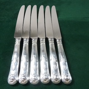 Op de afbeelding: Set van zes zilveren messen met sierlijke, bloemmotief handvatten en glanzende bladen. De messen zijn gerangschikt op een donkergroen oppervlak, wat hun elegante ontwerp benadrukt.