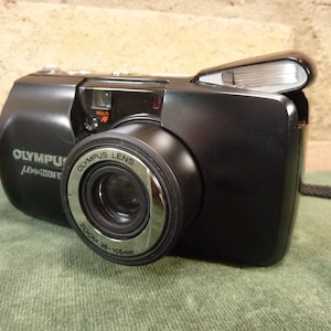 May include: A black Olympus mju zoom 105 camera with a silver lens ring. The lens has the text "Olympus Lens Zoom 38-105mm" printed on it. The camera has a built-in flash.