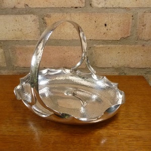 May include: A silver serving basket with a hammered finish and a decorative scalloped edge. The basket has a single handle and is oval shaped.