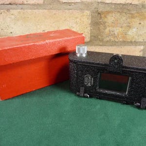 May include: A black camera with a silver lens and two silver knobs on the top. The camera is in a red box.