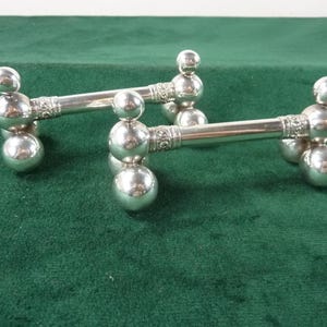 May include: Two silver knife rests. Each rest features a central bar with decorative detailing, supported by three spherical feet. The bar ends have a larger sphere and a smaller sphere. The rests are on a green surface.