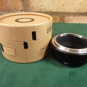 Puede incluir: Un adaptador de lente negro con detalles plateados. El adaptador tiene "FD-E" impreso en el lateral. El adaptador está sobre una superficie verde junto a una caja de cartón marrón con el texto "Lens Mount Adapter" impreso en el lateral.