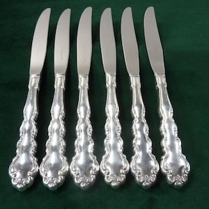 Könnte beinhalten: Sechs silberne Buttermesser mit verzierten, floralen Griffen. Die Klingen sind gebogen und glatt. Die Messer sind auf einer dunkelgrünen Oberfläche angeordnet, was ihr elegantes Design hervorhebt. Die Messer sind etwa 20 cm lang.