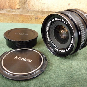 May include: A black Konica Hexanon AR 28mm f/3.5 lens with a silver Konica lens cap and a black Konica lens cap.