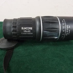 May include: A black Suncore monocular with a lens cap and strap. The monocular has the text "SUNCORE 10x40 114m/1000m" printed on the side. It also has a day and night field of view indicator.