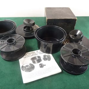 May include: A set of black film developing equipment, including tanks, reels, and a box. The items are arranged on a green surface, with a printed instruction sheet visible. The box has the text "PATERSON Tank".