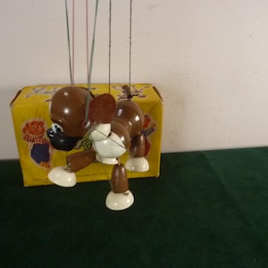 May include: A brown and white wooden dog marionette with a black nose and ears, suspended by thin strings. The puppet is positioned in front of a yellow cardboard box featuring illustrated characters and text, including the word 'Puppet'.