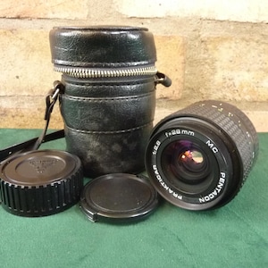 May include: A black Praktica Pentacon 28mm camera lens with a black leather case, lens cap, and lens cover. The lens has the text "f=28mm 1:2.8 MC Pentacon Praktica" printed on it. The case has a zipper closure and a strap.