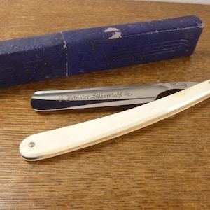 May include: A straight razor with an off-white handle and a metallic blade featuring the engraved text Feinster Silberstahl. The razor is accompanied by a rectangular, dark blue textured storage case with visible wear on the top surface.