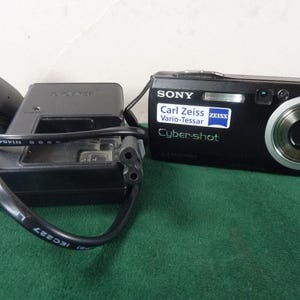 May include: A black Sony Cybershot digital camera with a Carl Zeiss Vario-Tessar lens, next to a black charger and power cord. The camera has a silver lens ring and a flash. The charger has a black power cord.