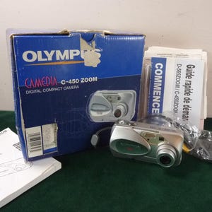 Könnte beinhalten: Eine Olympus Camedia C-450 Zoom digitale Kompaktkamera mit Originalverpackung und Handbüchern. Die Box ist blau mit weißem Text und einem Bild der Kamera. Die Kamera ist silber und schwarz.