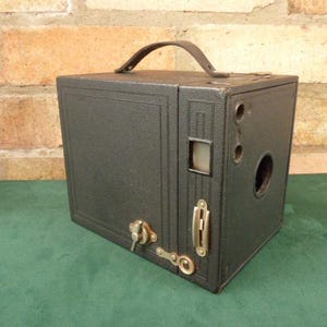 May include: A black vintage box camera with a leather carrying handle. The camera has a viewfinder, a lens, and a shutter release button.